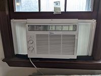 Full frontal view of the Midea window air conditioner installed in a window frame, showing vents, control dials, and side panels.