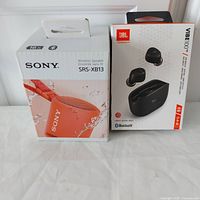 Photo showing boxed Sony SRS-XB13 speaker and boxed JBL Vibe 100TWS earbuds side by side.
