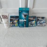 Full lot of men's shaving products shown in single image including Bulldog shave gel, Gillette Mach 3 razor handle with cartridges, and three packages of replacement cartridges in varied counts and types.
