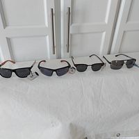 Four pairs of men's sunglasses on white cloth background, showing variety of styles and tags attached.