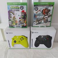 Two new sealed Xbox One games Skate 3 and Plants vs. Zombies Garden Warfare, with two Xbox wireless controllers in Electric Volt and Carbon Black colors in boxes in front.