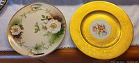 Two decorative plates side by side showing front designs, one with yellow-gold textured border and floral center, the other with cream background and white flowers.