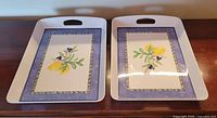 Top view of two large rectangular plastic serving trays with floral and fruit design and built-in handles.