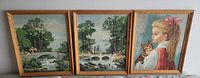 Three framed vintage paint by numbers paintings hung side by side. Two are landscape scenes with bridges over water and trees, the third is a portrait of a girl holding a cat.
