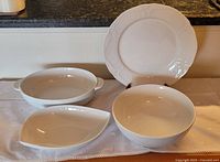 Four white ceramic serving dishes displayed on a table with a white cloth, including an ornate round plate, plain large bowl, oval handled dish, and leaf-shaped serving dish.