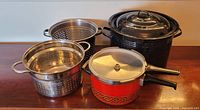 Photo of the lot showing the black speckled enamel lobster cooker, two stainless steel strainers, and the red pressure cooker with lid and handles.