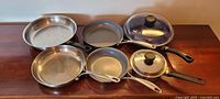 Photo of various stainless steel and cast iron pans and saucepans with glass lids, showing different sizes and handle types on a wooden surface