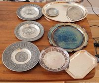 Overview of eight assorted pewter, silver plate, and chrome plates and trays displayed on wooden surface. Includes Canadian provinces-themed and anniversary collectibles.