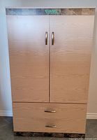 Front view of pantry storage cabinet closed, showing two vertical doors and two drawers below with silver handles, light wood grain finish, marble-look trim at top.