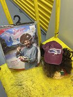 Sealed Bob Ross costume kit and Forrest Gump cap and wig on yellow surface