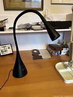 Black LED desk lamp with gooseneck on wooden desk, showing its conical base and lamp head, along with some background desk items.