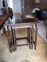Set of three dark brown wooden nesting tables arranged nested with visible turned spindle legs, photographed in room with mid-century furniture.