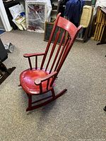 Side view of the red wooden rocking chair showing the curved armrests, seat, and rockers with visible wear.