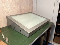 View of light table angled showing metal frame and frosted glass surface on green tabletop with wall background.
