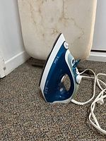 T-fal blue and white corded steam iron on carpet next to ironing board.