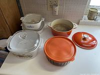 Overview of orange enameled cast iron Dutch oven with lid, white and blue floral Corningware casserole dishes with lids, and round orange casserole dish with lid on counter.