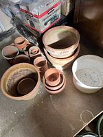 Multiple gardening clay pots of various sizes grouped with one white ceramic pot, all showing signs of prior outdoor use such as dirt and moss stains.