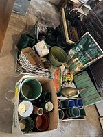 Wide view of assorted gardening supplies including pots, watering cans, trays, bags of seed and fertilizer, garden edge fencing, and other items on floor and table.