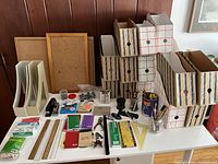 Photo showing assortment of office supplies spread on a table including file boxes, cork boards, notebooks, staplers, rulers, pencils, and containers with drawing and writing materials.