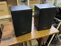 Pair of black Realistic Minimus 11 speakers on wooden surface showing front metal grills with logo