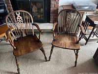 Two wooden captain's chairs shown side by side on carpet. Both chairs have spindle backs with carved central splats, curved armrests, turned legs, and stretchers for stability. Dark wood stain with visible grain.