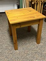Small square wooden table made from knotty pine wood with natural wood grain and knots, straight legs, placed on carpeted floor.