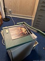Paper cutter sitting on a rectangular off-white cabinet, with green grid base, metal cutting arm extended to the side, and a photograph partially under the blade ready for cutting.