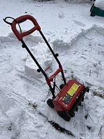 Red electric snow thrower standing on snow, showing entire unit with handle, wheels, and front clearing blades.