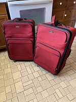 Two large red fabric Ricardo Beverly Hills suitcases standing upright on a tiled floor in a kitchen environment. Both suitcases have black zippers, exterior pockets, and fabric handles on the top and side.