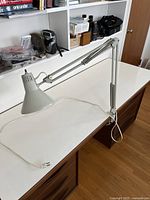 Gray/white metal desk lamp with adjustable arm clamped to edge of wooden desk showing overall lamp with cord and plug on desk surface.