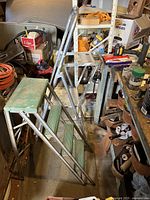 Photo showing two small step ladders placed side by side in a cluttered workshop or storage area. One ladder is aluminum with a handle and paint tray, the other is wood and metal with green steps and wooden top.