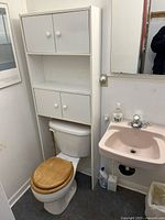 View of assembled white over-the-toilet cabinet over a toilet with two cabinets with doors and an open middle shelf, white painted MDF structure, situated next to a sink and mirror.
