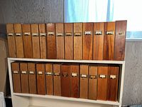 Two rows of 25 vintage wooden file boxes arranged on shelves, showing wood grain, metal label-holder pulls, varying wood tones and some wear.