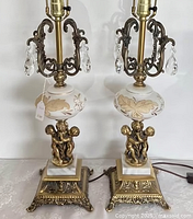 Front view of the pair of vintage brass and glass lamps with cherub bases, showing ornate bases and glass midsection with leaf motifs.