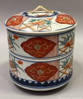 Front angled view of the antique Japanese porcelain jubako bento box showing two stacked tiers with red floral and geometric painted designs, blue patterned border at base, and matching lid with handle.