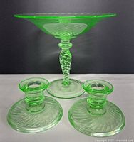 Front view of green uranium glass footed candy dish and two candle holders on white and black background, showing the swirl pattern and twisted stem on candy dish