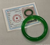 Photo shows the jade bangle alongside its certification card detailing identification and material authenticity.