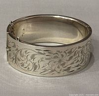 Front angled view showing wide bangle with etched floral pattern and safety chain clasp.