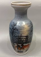 Front and partial side view of the vase showing hand painted winter landscape, two people and a cow in snow with blue sky background and gold rim detail.