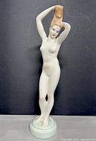 Full front view of the white porcelain figurine of a nude female figure holding a brown object above her head, standing on a round base.