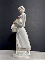 Side view of the porcelain figurine of a woman holding a chicken and a basket, standing against a black background showing the full figure and base.
