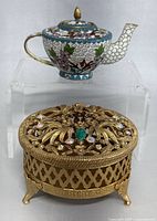 Photo of the small cloisonne teapot sitting behind the gold-tone metal filigree trinket box on a clear acrylic stand.