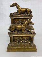 Front view of one brass dog bookend showing reclining dog and embossed decorative base