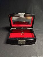 Jewelry box open showing red velvet interior and single ring inside with metal clasp visible on front.