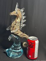 Side view of Dale Tiffany art glass seahorse with clear glass fins and spines, black eyes, and swirling amber and brown body, mounted on blue glass wave base with Coca-Cola can for size reference.