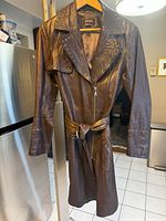 Full view of the dark brown leather trench coat hanging on a wooden hanger showing zippered front, belted waist, and metal grommet trims on collar and sleeves.