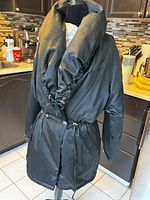 Full front view of black Hilary Radley puffer coat on dress form in kitchen, showing oversized ruched collar, adjustable drawstring waist, side pockets, and button closure.