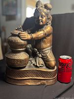 Front view of a copper-colored cast metal statue showing Baby Krishna kneeling and balancing stacked jars, with a Coca-Cola can for size reference about 15.5 inches tall