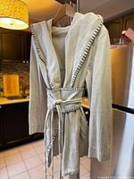 Full view of beige hooded wrap coat on a hanger in a kitchen showing the belted waist and hood with contrast black stitching.