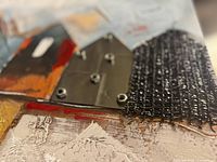 Close-up of textured mixed media elements including painted canvas, metal screws, and mesh metal piece enhancing 3D effect on abstract houses.
